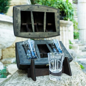 Puede incluir: Una caja de madera oscura con tres vasos de chupito grabados con el nombre "DANIEL". La caja está abierta, mostrando los vasos y sus compartimentos. La caja está colocada sobre una superficie de piedra, con un fondo borroso.