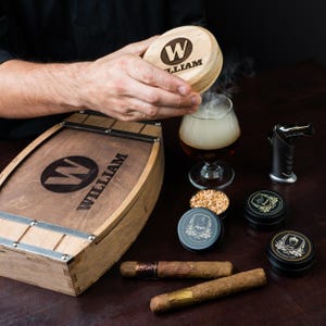 Può includere: Una scatola di legno a forma di botte con il nome "WILLIAM" e un logo "W", un bicchiere di liquido con fumo, sigari e piccole lattine. È presente anche un piccolo accendino a torcia. Il coperchio della scatola viene sollevato.