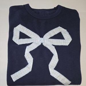 Puede incluir: Sudadera azul marino con un gran lazo de vichy azul claro. El lazo está centrado en la parte delantera y tiene un borde festoneado. La sudadera está doblada cuidadosamente.