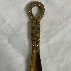 May include: A vintage brass shoe horn with a twisted handle. The horn has a long, slender shape with a curved end for easy shoe insertion. The brass has a warm, golden hue, and the handle is intricately designed.