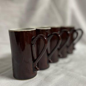 May include: A set of five brown ceramic mugs with a glossy finish. Each mug has a curved handle and a white rim. The mugs are arranged in a row, with the first mug in focus.