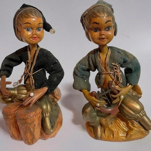 May include: Two vintage figurines of musicians. Each figure has a wooden head with painted features, wearing fabric clothing. One figure is playing drums, the other a stringed instrument. The figures are seated on a gold-colored base.