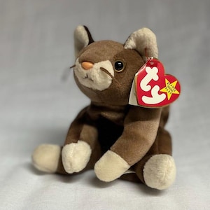 May include: A brown and white plush cat toy with a red heart-shaped tag. The cat has a light brown face, ears, and paws, with dark brown markings. The tag reads "Beanie Baby" and "Original Baby".