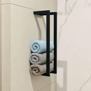 Könnte beinhalten: Ein schwarzer Metall-Handtuchhalter an einer hellbeige gefliesten Wand, der drei gerollte Handtücher hält. Die Handtücher sind hellblau, grau und beige. Der Halter hat ein minimalistisches, modernes Design mit einem rechteckigen Rahmen.