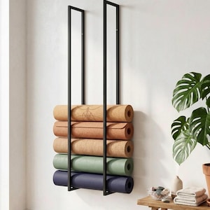 Puede incluir: Un estante de pared de metal negro que sostiene cinco esterillas de yoga enrolladas en varios colores, incluyendo bronceado, marrón claro, beige, verde salvia y azul oscuro. El estante cuenta con dos marcos verticales para las esterillas.