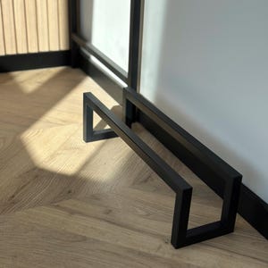 May include: A black metal towel rack with a geometric design. The rack is made of square tubing and has a minimalist style. The rack is positioned against a white wall and a wooden door.