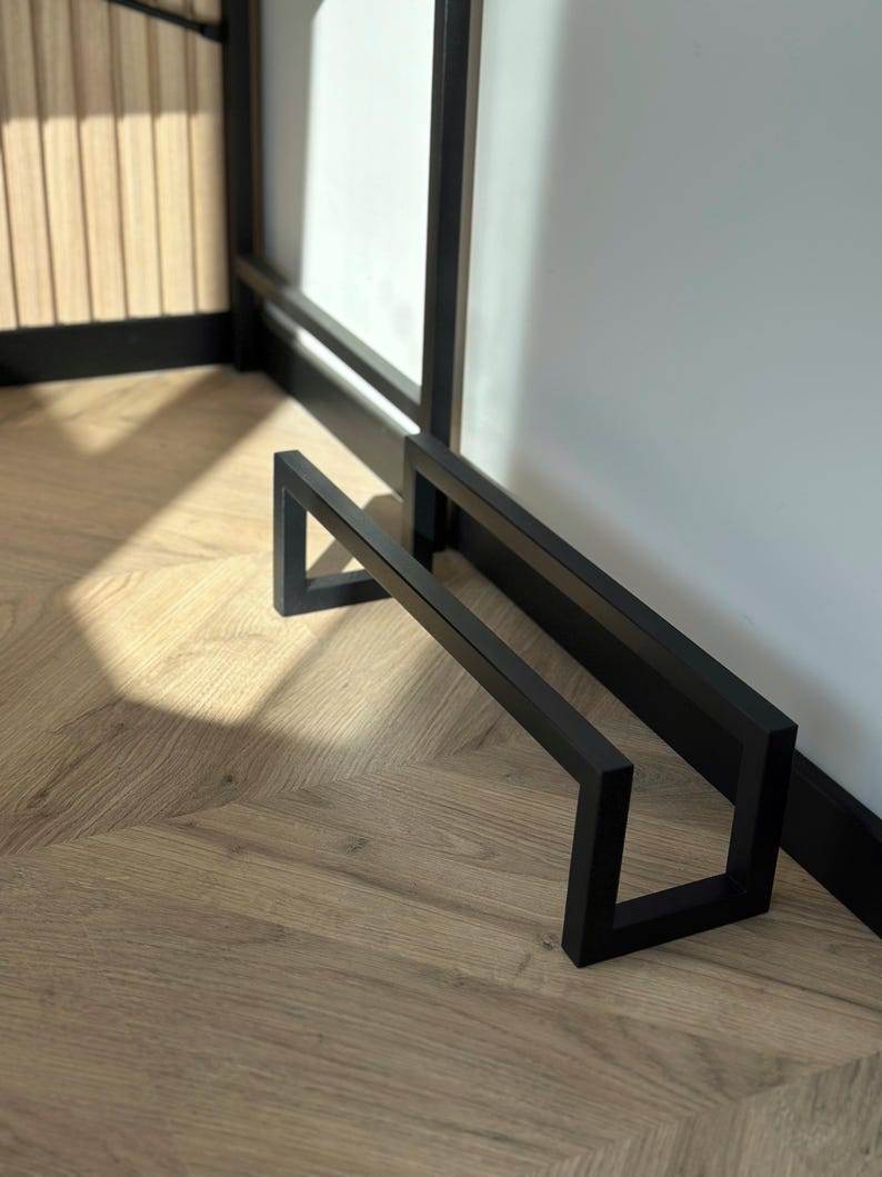May include: A black metal towel rack with a geometric design. The rack is made of square tubing and has a minimalist style. The rack is positioned against a white wall and a wooden door.