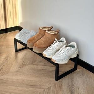 Puede incluir: Un zapatero de metal negro que exhibe cuatro pares de zapatos. Los zapatos incluyen zapatillas de color azul claro, botas marrones con plataforma y forro beige, y dos pares de zapatillas blancas. El zapatero está sobre un suelo de madera.