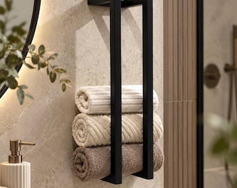 May include: A matte black metal wall-mounted towel rack featuring a vertical rectangular frame design. The rack holds three neatly rolled towels in stacked order, with the top towel being white, the middle beige, and the bottom light brown.