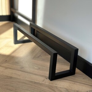 May include: A black metal shoe rack with a rectangular design. The rack is made of thick, square metal bars and is positioned against a white wall and a wooden floor. The rack is approximately 24 inches long and 12 inches high.