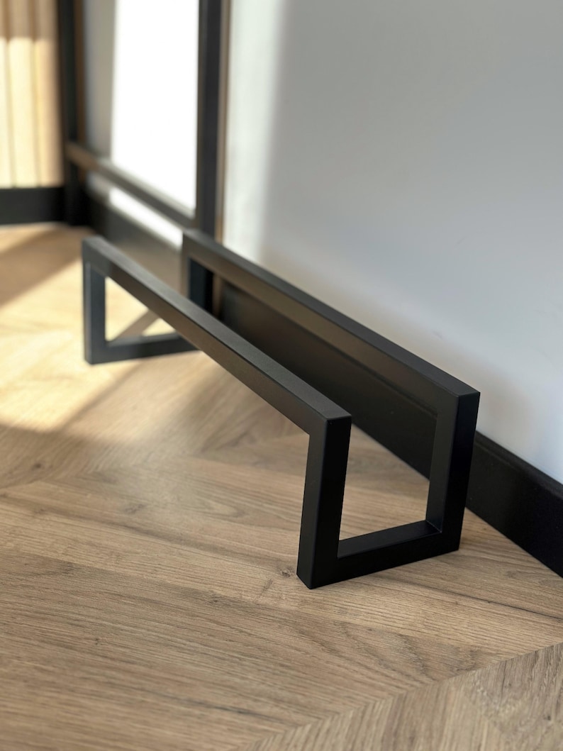May include: A black metal shoe rack with a rectangular design. The rack is made of thick, square metal bars and is positioned against a white wall and a wooden floor. The rack is approximately 24 inches long and 12 inches high.