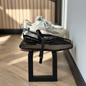 May include: Black mesh flats with a strap and buckle detail, displayed on a black square stand. A pair of white and grey sneakers with green accents are in the background. The shoes are on a wooden floor.