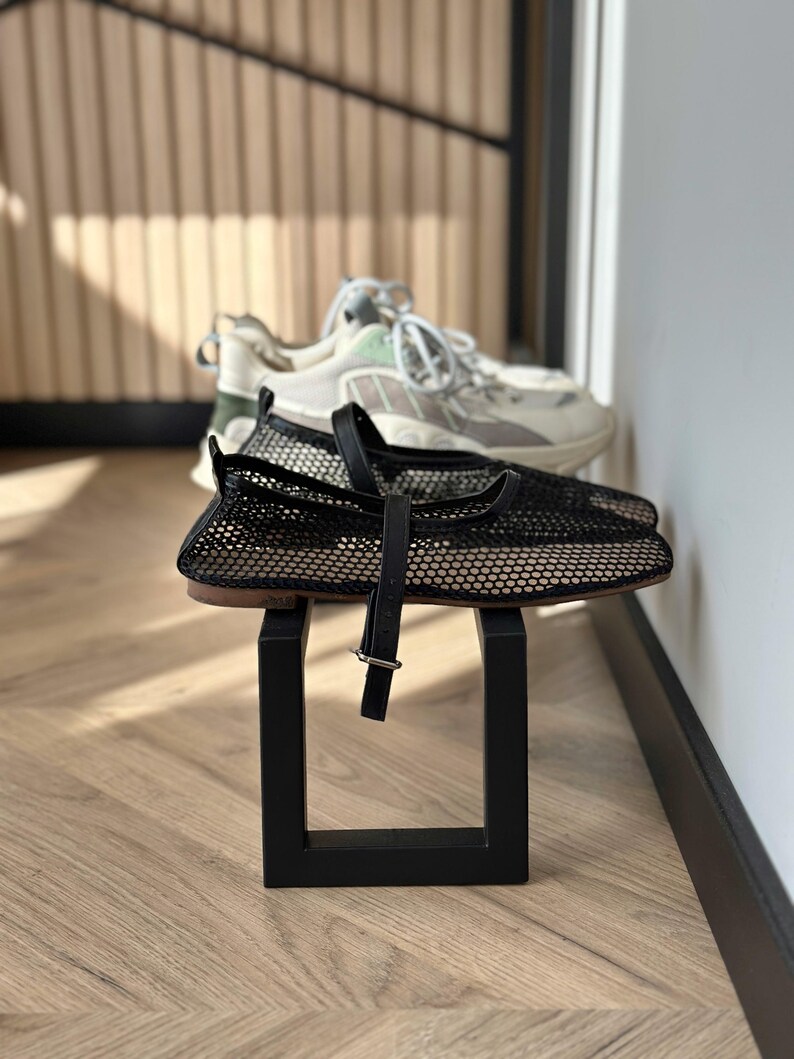 May include: Black mesh flats with a strap and buckle detail, displayed on a black square stand. A pair of white and grey sneakers with green accents are in the background. The shoes are on a wooden floor.