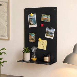 May include: A rectangular black metal wall-mounted magnetic board with an integrated shelf. The board displays two landscape photographs, two rectangular magnets labeled LISBOA and BUDAPEST, and a yellow sticky note with the text Don't forget to be awesome.