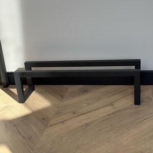 May include: Two black metal stands with a rectangular design. The stands are placed on a wooden floor with a light brown color. The stands are against a white wall.