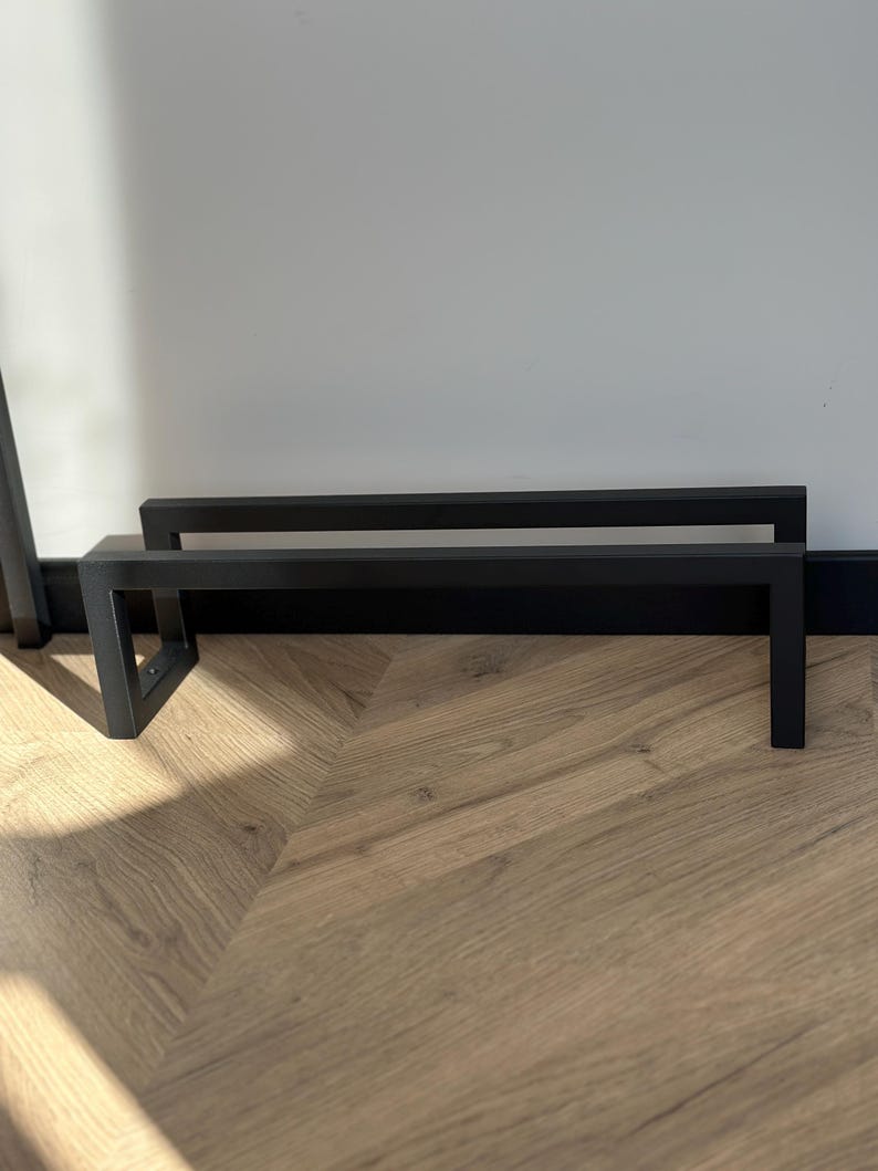 May include: Two black metal stands with a rectangular design. The stands are placed on a wooden floor with a light brown color. The stands are against a white wall.