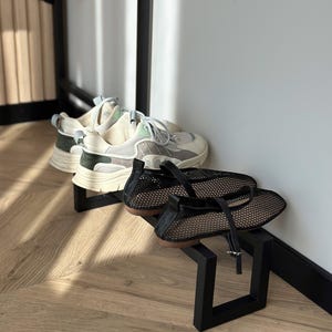 Puede incluir: Dos pares de zapatos exhibidos en soportes de metal negro. El par superior es una zapatilla gruesa con detalles crema, grises y verdes. Debajo, un par de bailarinas de malla negra con correa y hebilla.