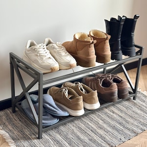 Puede incluir: Un zapatero de metal gris con tres estantes, que contiene varios zapatos. El estante superior muestra zapatillas blancas, botas UGG color canela y botines negros. El estante del medio tiene zapatillas marrones. Hay zapatillas azules en el estante inferior.