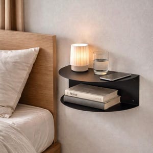 Puede incluir: Una estantería negra de dos niveles, montada en la pared, con un diseño curvo. La balda superior contiene una pequeña lámpara, un vaso de agua y un smartphone. La balda inferior muestra dos libros. La estantería está montada en una pared de color claro.
