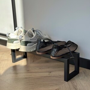 May include: A black metal shoe rack holding two pairs of shoes. One pair is white and gray sneakers with green accents. The other pair is black mesh flats with a buckle. The rack is on a wooden floor.