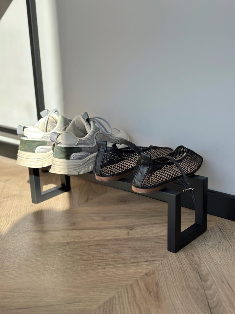 May include: A black metal shoe rack holding two pairs of shoes. One pair is white and gray sneakers with green accents. The other pair is black mesh flats with a buckle. The rack is on a wooden floor.