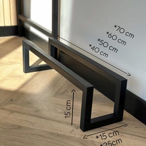 May include: Black metal shelf brackets with dimensions of 15 cm x 25 cm and heights of 40 cm, 50 cm, 60 cm, and 70 cm. The brackets are displayed on a wooden floor, showcasing their modern design.