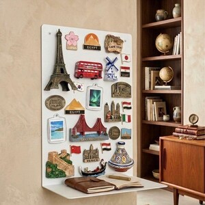 May include: A white magnetic board displaying travel-themed souvenir magnets. The magnets depict landmarks like the Eiffel Tower, Golden Gate Bridge, and a London double-decker bus. A brown leather journal and pen rest on a shelf below.