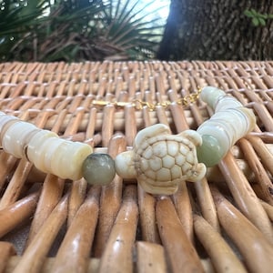 Pode incluir: Uma pulseira de contas com um pingente de tartaruga esculpida, contas de concha e detalhes em pedra verde. A pulseira é enfiada em uma corrente dourada e repousa sobre uma superfície tecida.