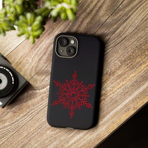 Peut inclure: Coque de smartphone noire avec un motif de flocon de neige rouge. L'étui est posé sur une surface en bois, avec un appareil photo et de la verdure en arrière-plan. Le flocon de neige est un motif symétrique détaillé.