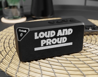 May include: A black rectangular Bluetooth speaker with the words "LOUD AND PROUD" in white. The speaker has control buttons on top and a mesh front. The speaker is on a woven mat.