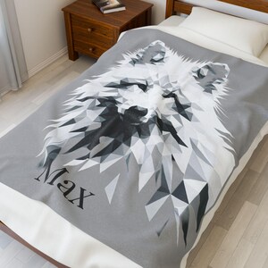 Op de afbeelding: Een grijze deken met een geometrisch ontwerp van een wolf in wit, grijs en zwart. De naam "Max" staat in het zwart onderaan. De deken ligt op een bed met een wit kussen.