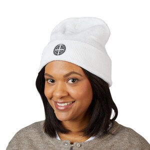 May include: A white beanie with a black circular emblem on the front. The emblem features a cross design with the text around the edge. The beanie is made of a ribbed knit material and is worn by a person.