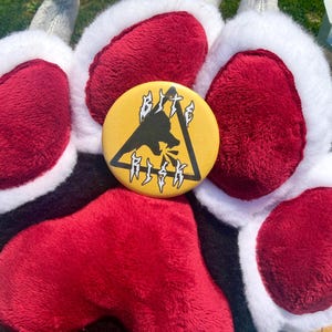 May include: A round yellow pinback button featuring a black silhouette of a wolf head inside a black triangle with the text BITE RISK in a jagged white font. The button is placed on a plush paw-shaped glove with red pads and white fur.