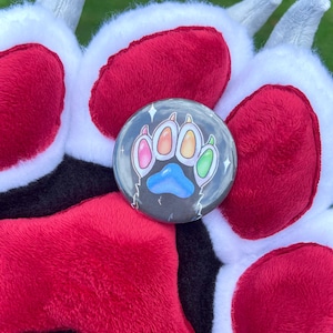 Op de afbeelding: Een ronde knoop met een kleurrijk pootafdrukontwerp, met roze, oranje, gele en groene tenen en een blauwe voetzool. De knoop is geplaatst tegen een pluche, harige achtergrond van rood, zwart en wit.