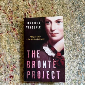 May include: A hardcover book titled "The Bronte Project" by Jennifer Vandever. The cover features a portrait of a woman and a quote from the New York Times Book Review. The book is set against a patterned background.