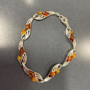 May include: Silver bracelet featuring leaf-shaped links adorned with amber-colored gemstones. The bracelet showcases a variety of amber hues, from light yellow to deep orange, creating a striking contrast against the silver setting. A delicate piece of jewelry.