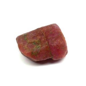 Könnte beinhalten: Ein roher, ungeschliffener Rubin-Edelstein mit tiefroter Farbe und grünen Flecken. Der Naturstein hat eine strukturierte Oberfläche und eine unregelmäßige Form, ideal für Schmuck oder Sammlungen.