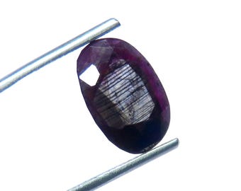 Könnte beinhalten: Ein facettierter, oval geformter, tiefvioletter Edelstein, der von einer silbernen Pinzette gehalten wird. Der Edelstein hat eine polierte Oberfläche mit inneren Streifen und eine helle, reflektierende Facette. Der Edelstein ist vor weißem Hintergrund.