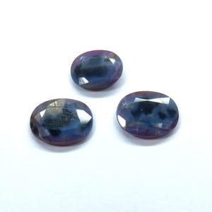 May include: Three oval-cut gemstones with a mix of blue, purple, and dark hues. The gems are faceted, reflecting light and creating a sparkling effect. The stones are set against a white background.