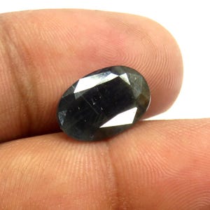 May include: A faceted, oval-shaped gemstone with a dark, smoky color. The gem has multiple facets that reflect light, creating a sparkling effect. The stone is held in a hand, showcasing its size and shape.