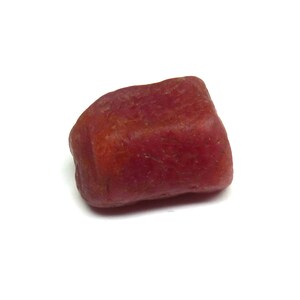 Puede incluir: Una gema lisa de color rojo púrpura con forma redondeada. La piedra tiene un aspecto natural, sin pulir, mostrando variaciones de color y textura. La gema se encuentra sobre un fondo blanco.
