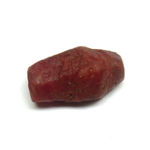 Könnte beinhalten: Ein rauer, ungeschliffener Rubin-Edelstein. Der Rubin hat eine tiefrote Farbe mit einer leicht strukturierten Oberfläche. Die Form ist länglich und unregelmäßig, mit abgerundeten Kanten. Der Edelstein ist vor einem weißen Hintergrund platziert.