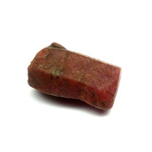 Puede incluir: Una gema de color marrón rojizo, de forma rectangular y de corte rugoso. La piedra tiene una superficie texturizada con toques de verde y un borde ligeramente redondeado. La gema está sobre un fondo blanco.