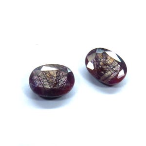 May include: Two oval-shaped gemstones with a deep burgundy hue and internal inclusions. The gems have a faceted cut, reflecting light and creating a sparkling effect. The stones are set against a white background, highlighting their color and clarity. These gemstones are suitable for jewelry making.