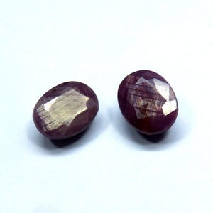 May include: Two oval-shaped, faceted gemstones with a deep red hue. The gems have a polished, reflective surface, with visible internal structures. The stones are set against a white background, highlighting their color and clarity. These gemstones are suitable for jewelry making.
