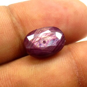 May include: A faceted, oval-shaped gemstone with a deep purplish-red hue. The gem has multiple facets that reflect light, creating bright white highlights. Internal inclusions create a unique pattern within the stone, adding to its visual interest. The gem is held in a hand.