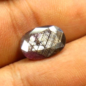 May include: An oval-shaped gemstone with a faceted surface. The gem has a mix of colors, including purple, gray, and black. The facets create a play of light and shadow, enhancing its brilliance. The gemstone is held in a hand.