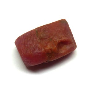 May include: A raw, reddish-brown gemstone with an uneven shape. The stone has a textured surface and varying shades of red and brown. The gemstone is set against a white background.