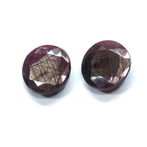 May include: Two oval-shaped, faceted gemstones in a deep reddish-purple colour. The gems have internal inclusions and a polished surface, reflecting light. The stones are set against a white background, highlighting their colour and clarity.