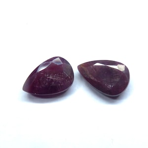 May include: Two faceted, pear-shaped gemstones with a deep red hue. The gems have a polished surface with visible internal structures and a bright, reflective facet. The gemstones are set against a white background.
